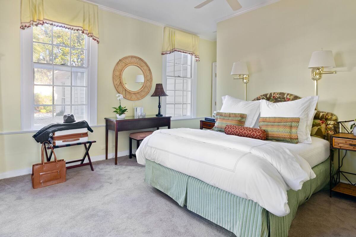 Une chambre à coucher comprenant un lit, des tables de chevet, un bureau, une chaise, un miroir et une fenêtre avec des rideaux.
