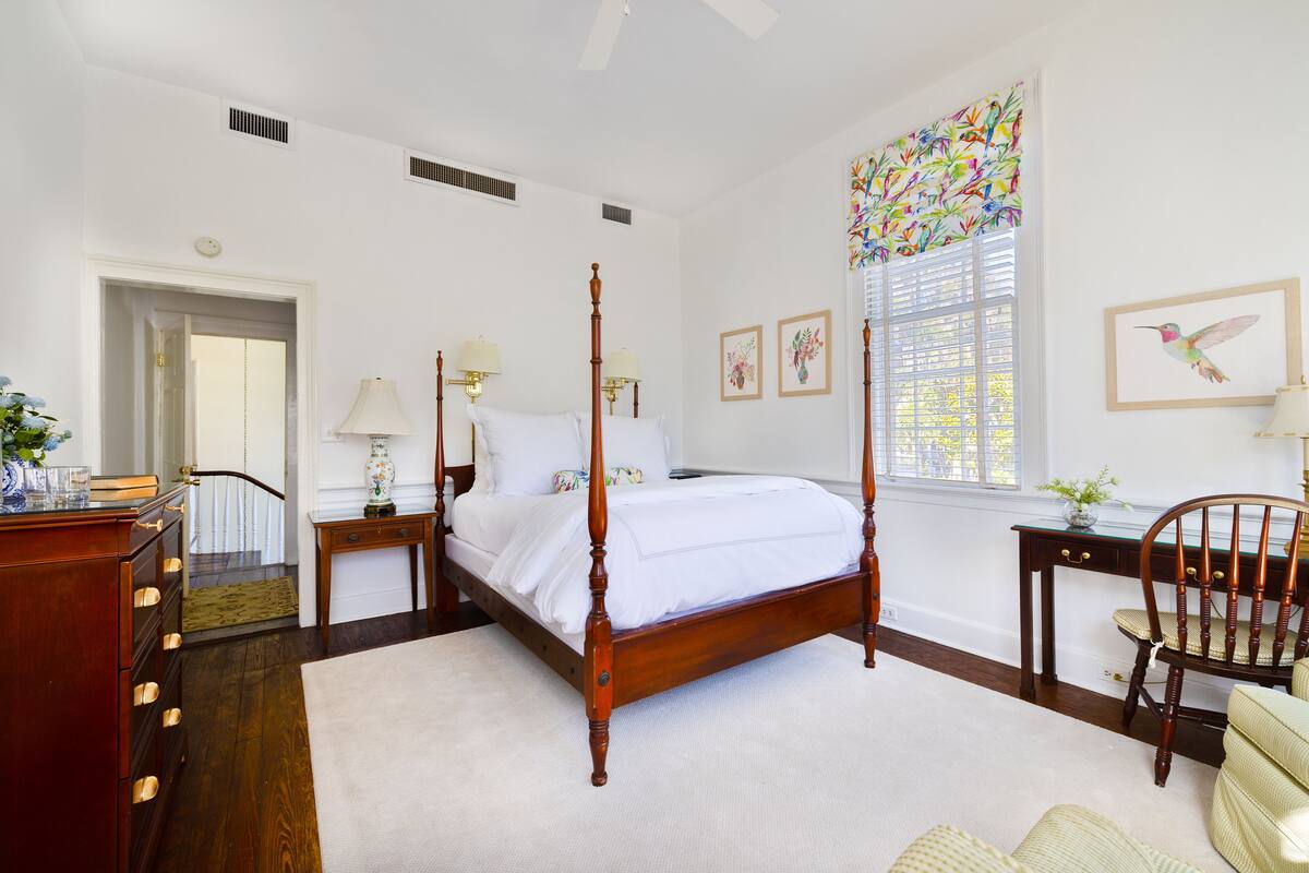 Une chambre à coucher avec un lit à baldaquin, une commode en bois, un bureau avec une chaise et une fenêtre donnant sur un paysage coloré.