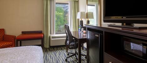 A hotel room with a bed, a desk with a television, a chair, a desk lamp, and a window with curtains.