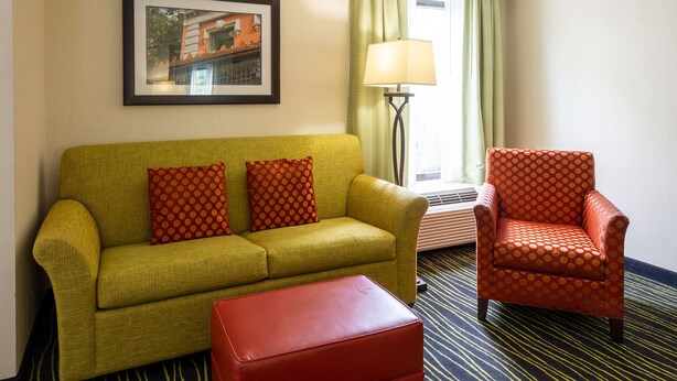Une chambre d’hôtel avec un canapé vert, des coussins à motifs rouges et orange, un pouf rouge et un fauteuil à motifs rouges. Un tableau encadré est accroché au mur, il y a une lampe sur pied et une fenêtre avec des rideaux verts.