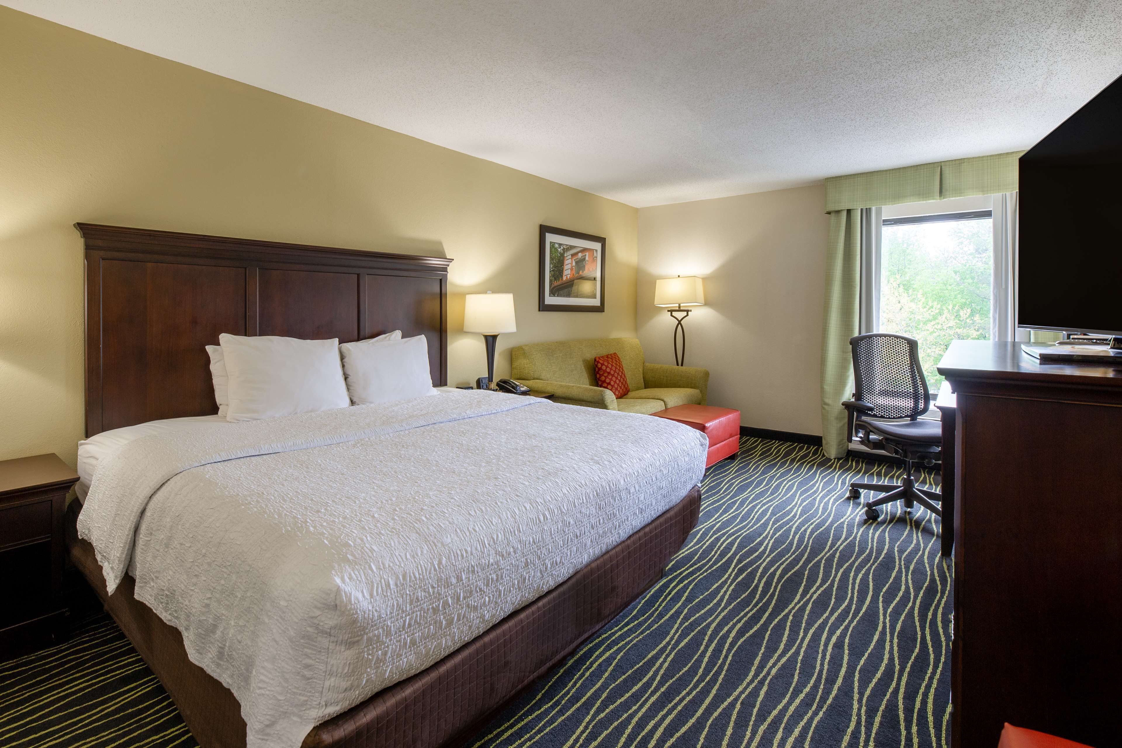 A hotel room with a large bed, a desk, a chair, a lamp, and a window.
