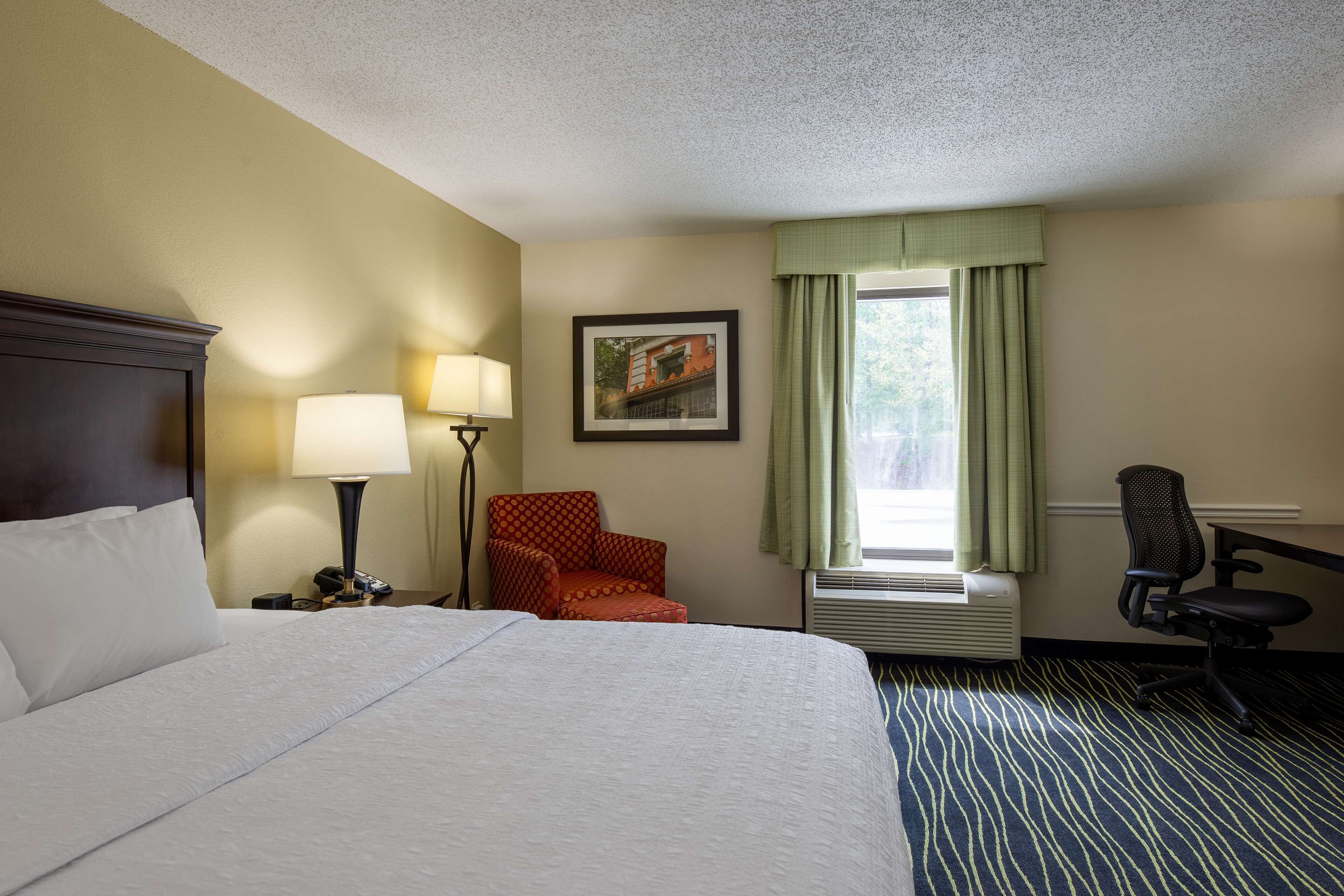 A hotel room with a large bed, a desk with a chair, a lamp, a framed picture on the wall, and a window with curtains.