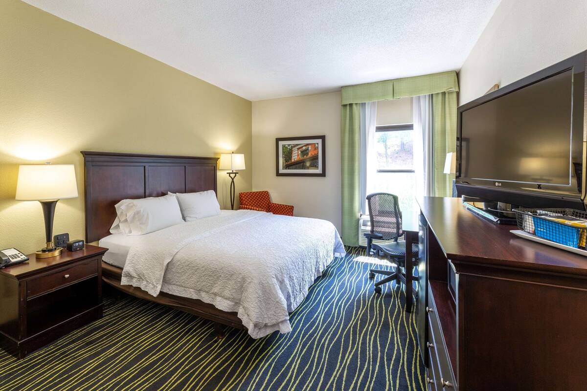 Habitación de hotel con cama, mesita de noche, escritorio, silla, televisor y ventana con cortinas.