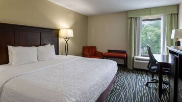 A hotel room with a large bed, a desk with a chair, a lamp, and a view of trees through the window.