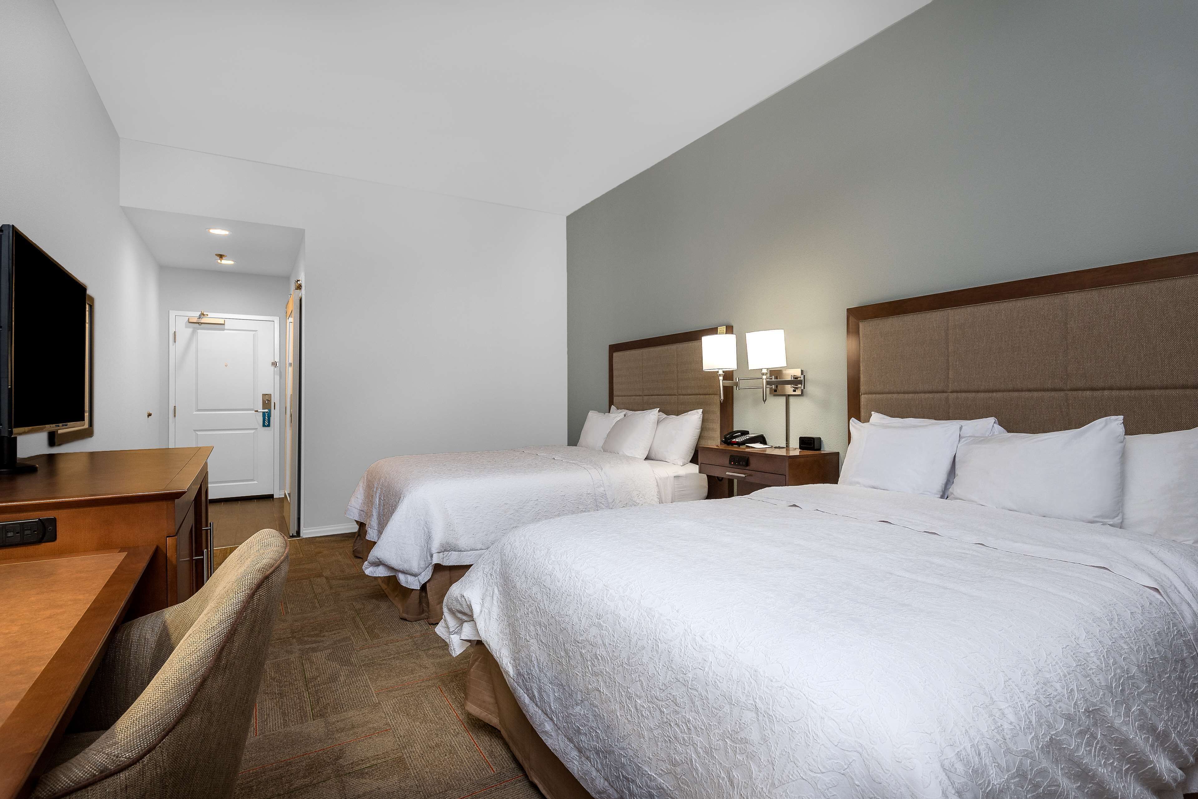A hotel room with two beds, a desk, a chair, a television, and a door.