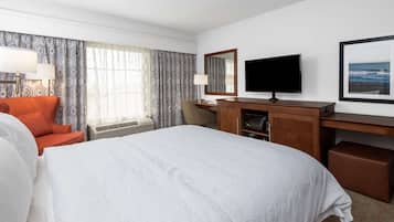 A hotel room with a large bed, a desk with a flat-screen TV, a chair, and a window with patterned curtains.