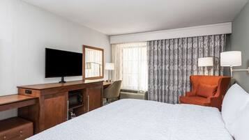 Habitación de hotel con una cama grande, un escritorio con televisor, una silla y una ventana con cortinas estampadas.