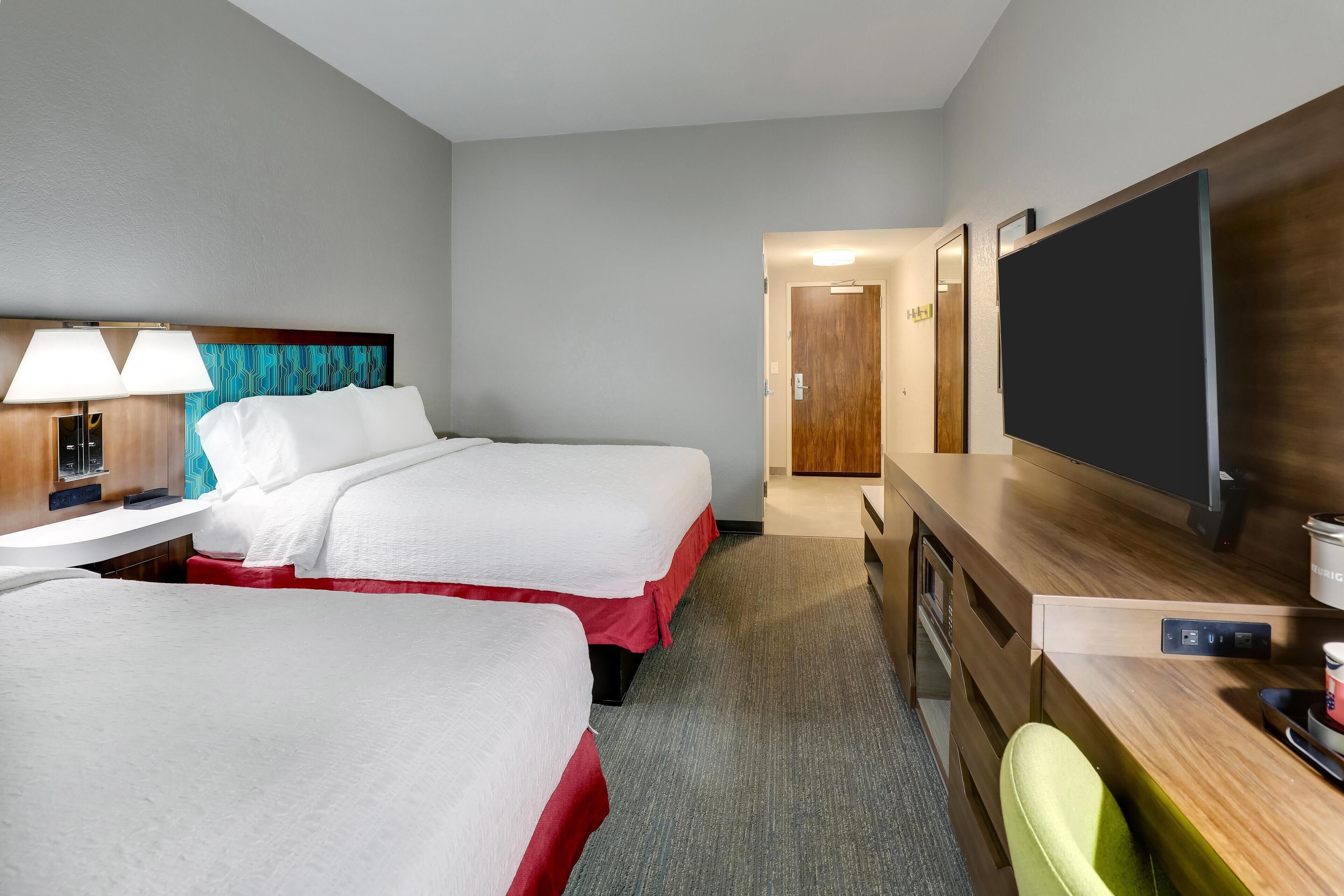 Habitación de hotel con dos camas, un escritorio, televisor de pantalla plana, una lámpara y cabecera de madera.