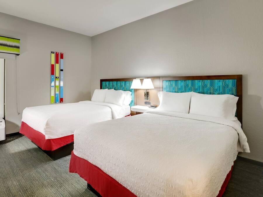 A hotel room with two beds, a window, an air conditioning unit, and a wall-mounted lamp.
