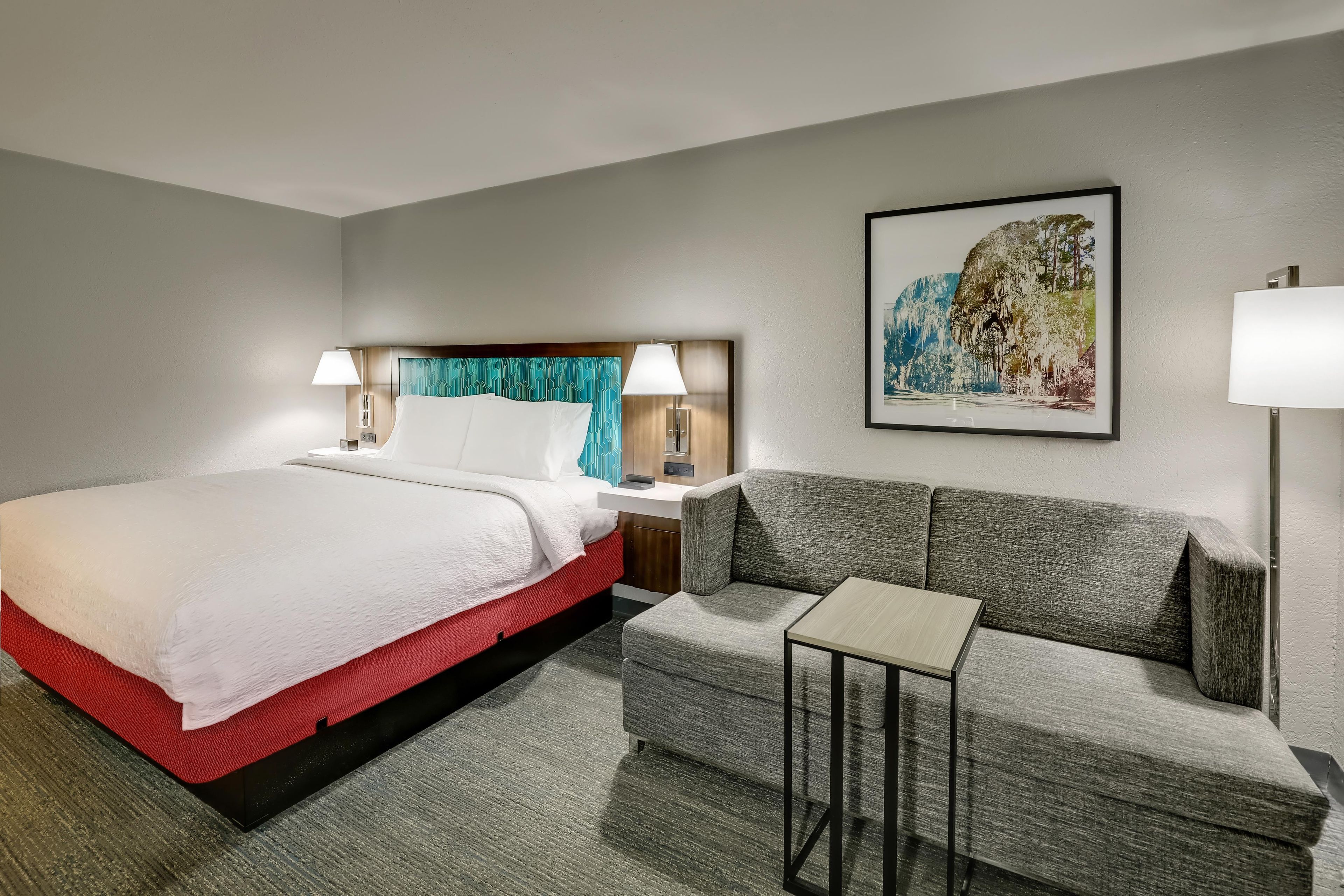 A hotel room with a bed, a sofa, a small table, and a painting on the wall.