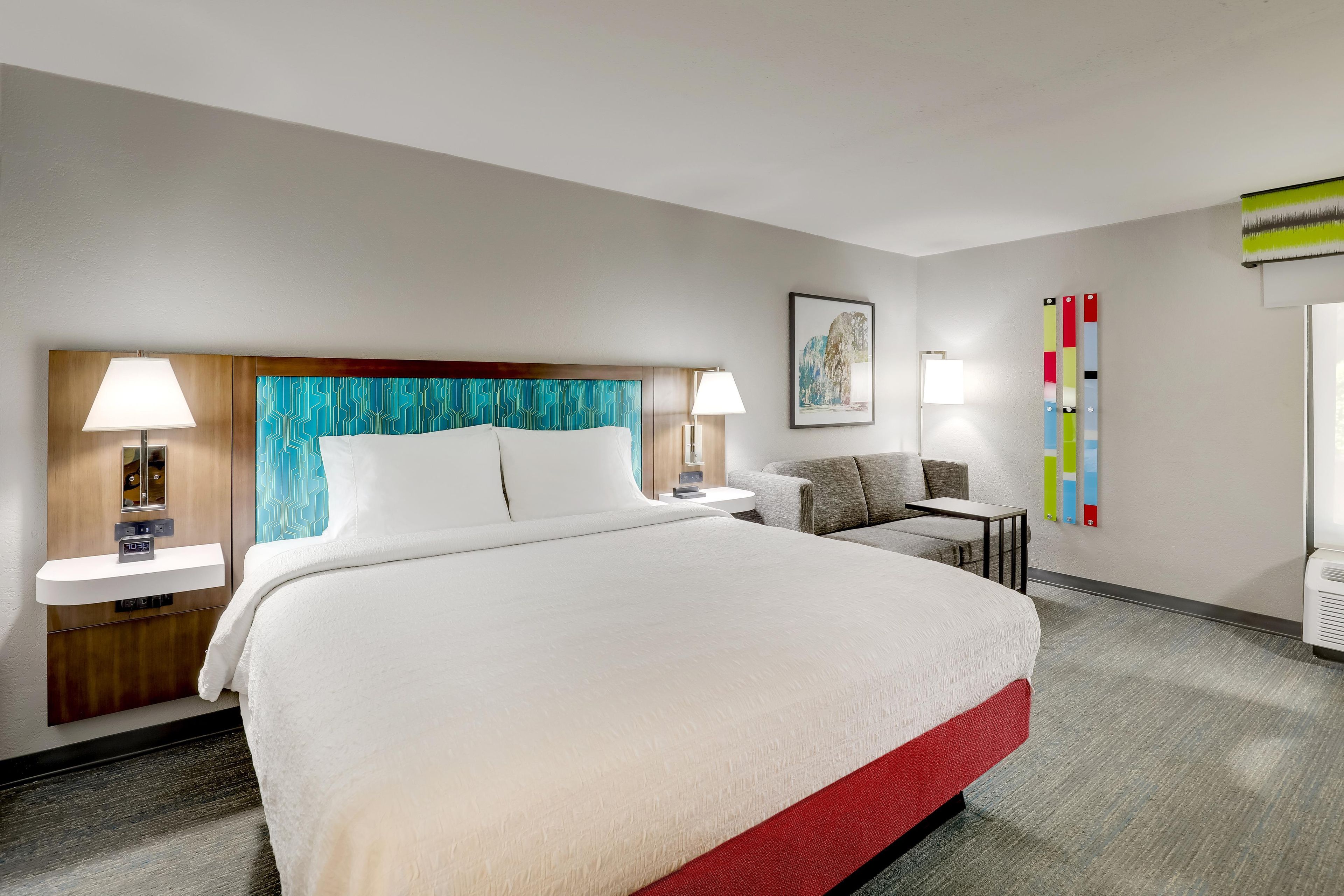 Une chambre d’hôtel avec un grand lit, deux tables de chevet, une chaise, un bureau et une œuvre d’art accrochée au mur.