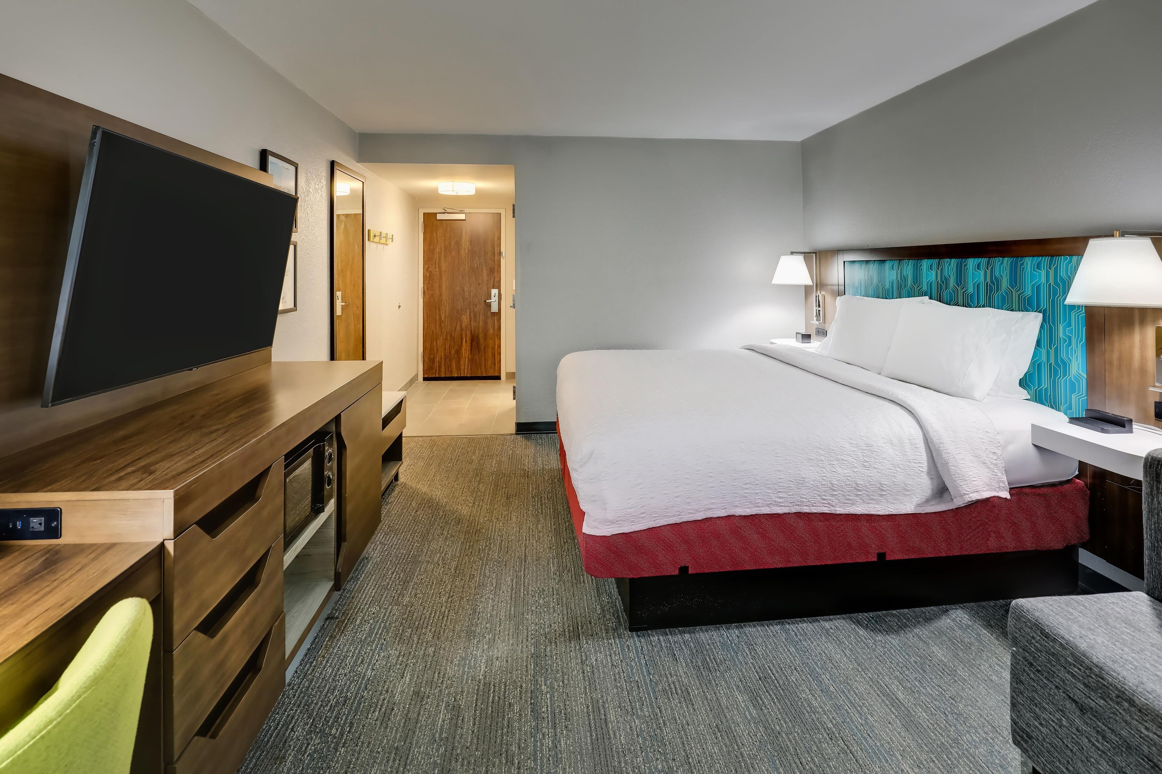 A hotel room with a bed, bedside table, lamp, TV, and a desk with a flat-screen TV.