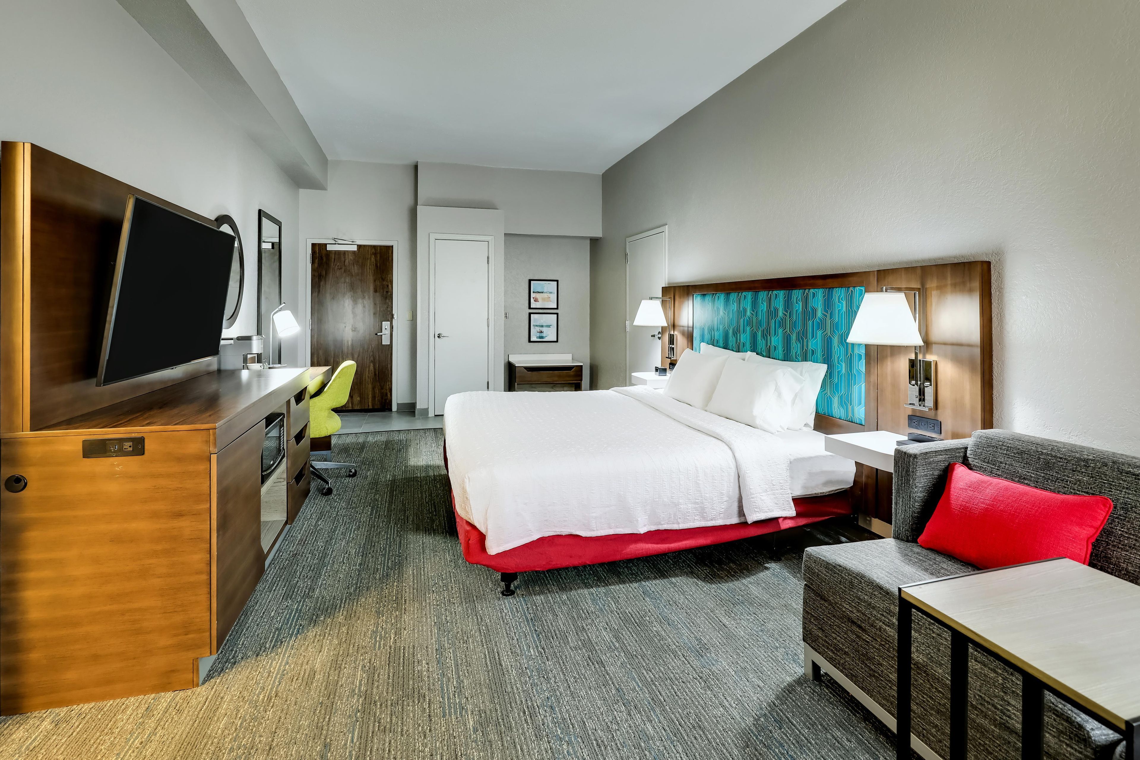 A hotel room with a bed, a desk, a chair, a TV, and a lamp.