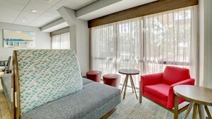 Una habitación de hotel moderna con un sofá gris, un sillón rojo, una mesita redonda y un ventanal con cortinas translúcidas.