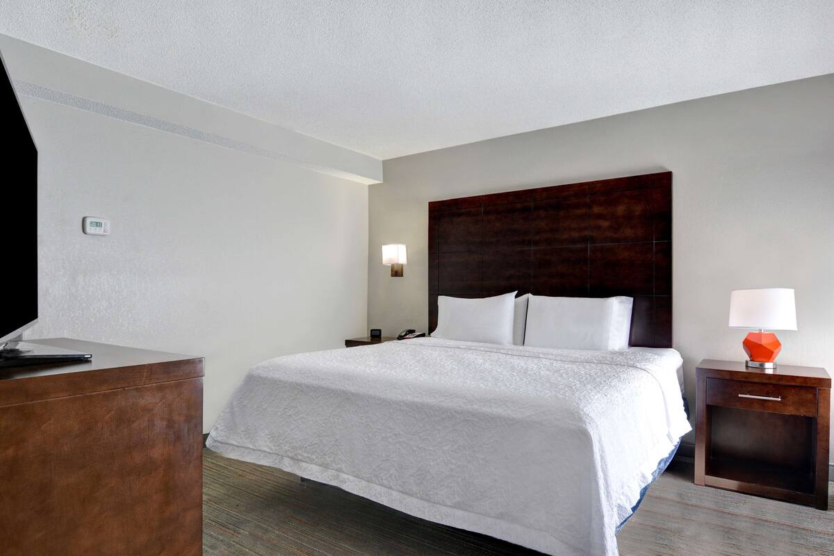 A hotel room with a large bed, a wooden dresser, a nightstand with a lamp, and a thermostat on the wall.