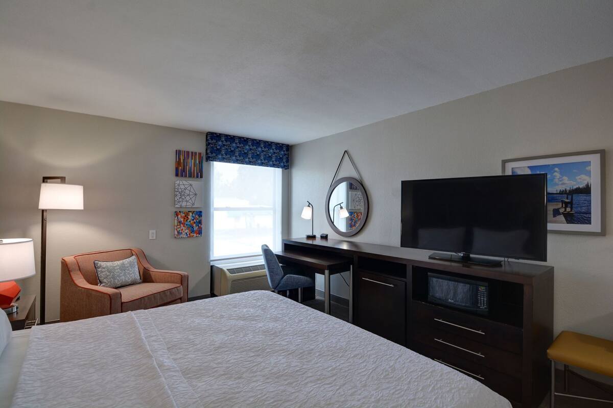 A hotel room with a bed, desk, TV, chair, and artwork on the walls.