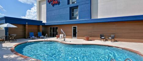 Una piscina con un edificio azul y blanco al fondo, un letrero que dice "Hampton Inn", y un área de patio con sillas y una mesa.