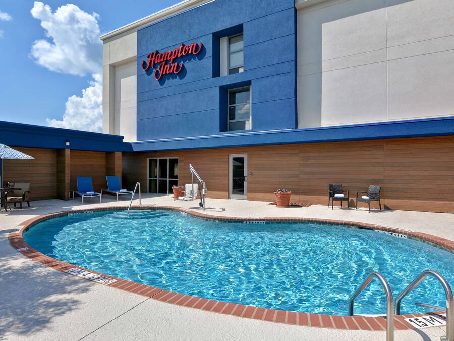 A swimming pool with a blue and white building in the background, a sign reading "Hampton Inn", and a patio area with chairs and a table.