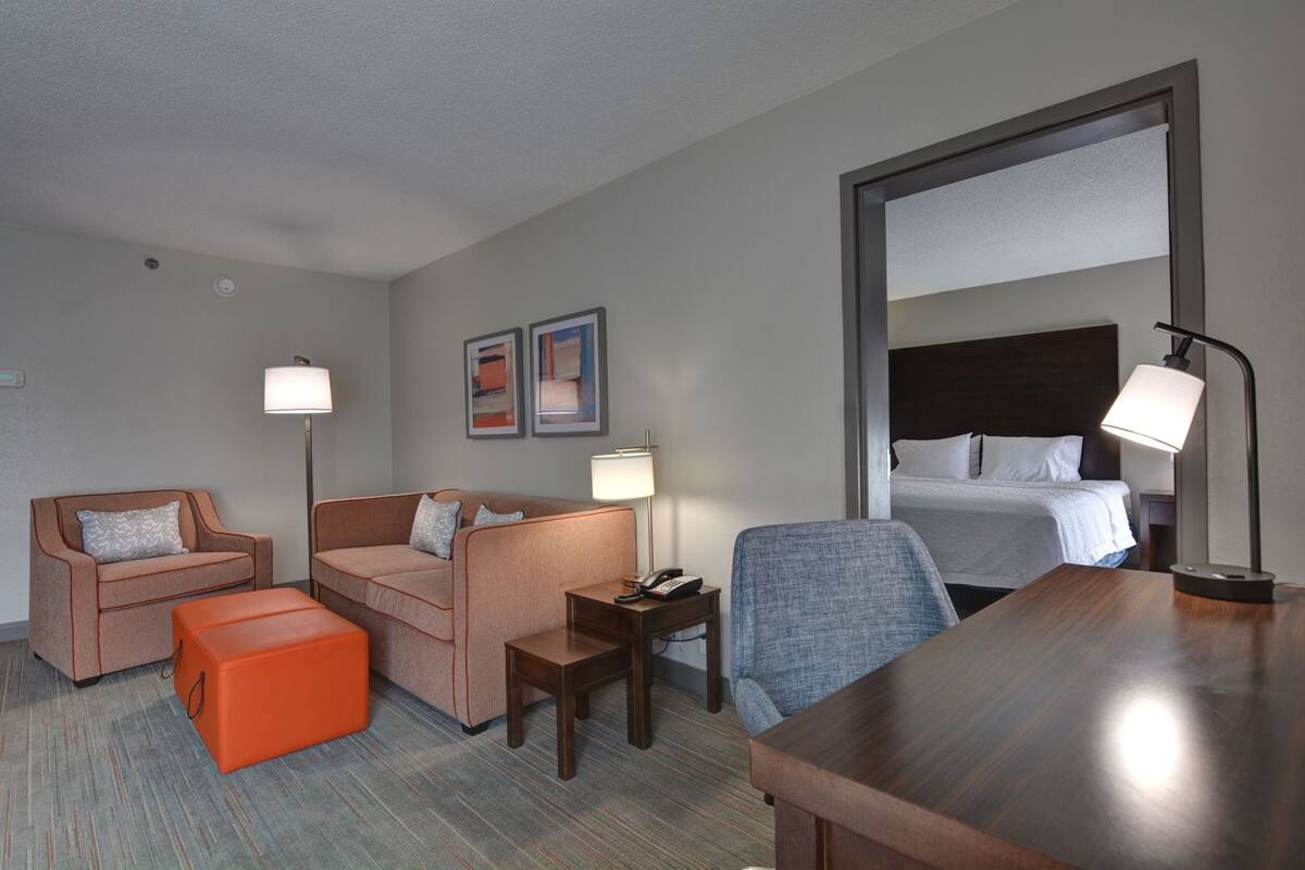A hotel room with two sofas, a small table, a chair, a wooden desk, a bedside lamp, a bed, and two framed pictures on the wall.