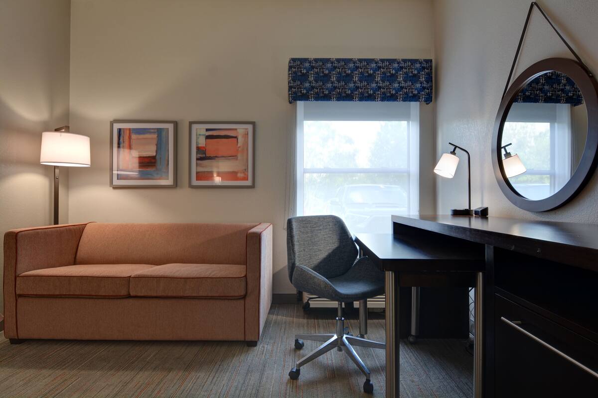 A room with a sofa, a desk, a chair, a lamp, and two framed artworks on the wall.