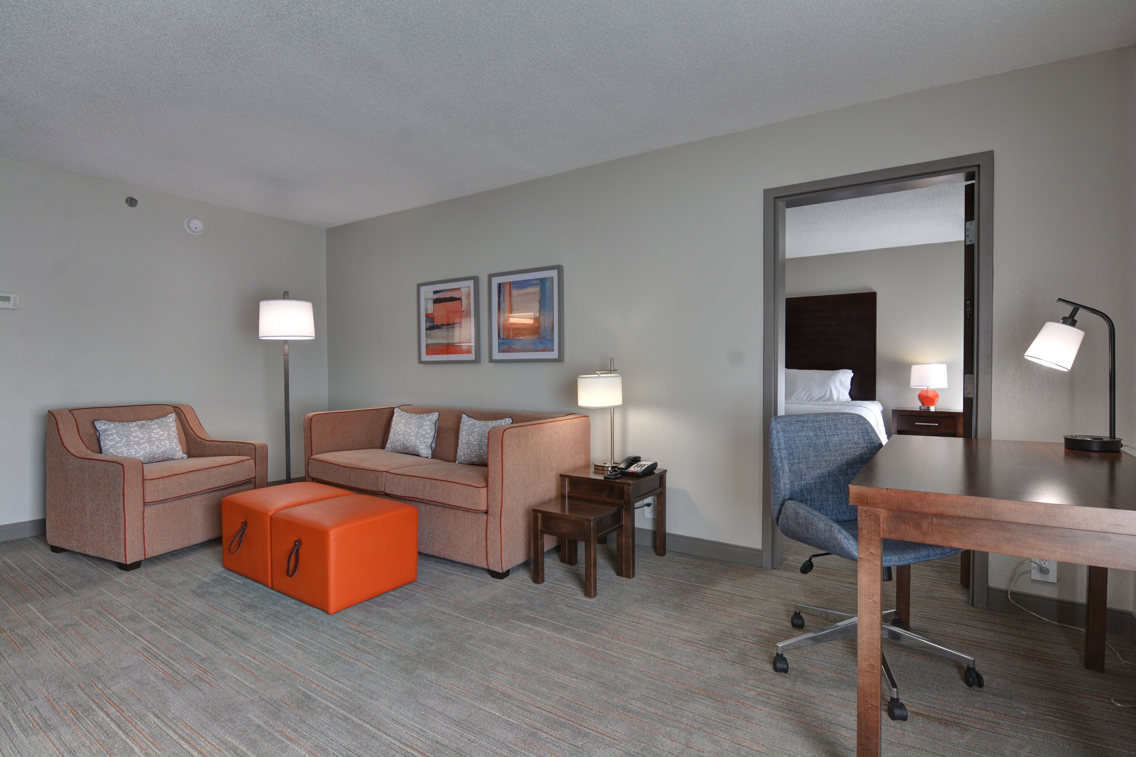 Una habitación de hotel moderna con dos sofás, un reposapiés naranja, un escritorio y una silla.