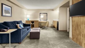 Habitación de hotel con sofá, escritorio, silla, reposapiés y televisor.