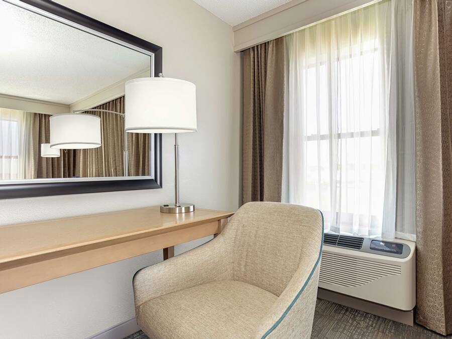 Une chambre d’hôtel avec un bureau en bois, un fauteuil beige, un grand miroir et une fenêtre avec des rideaux.