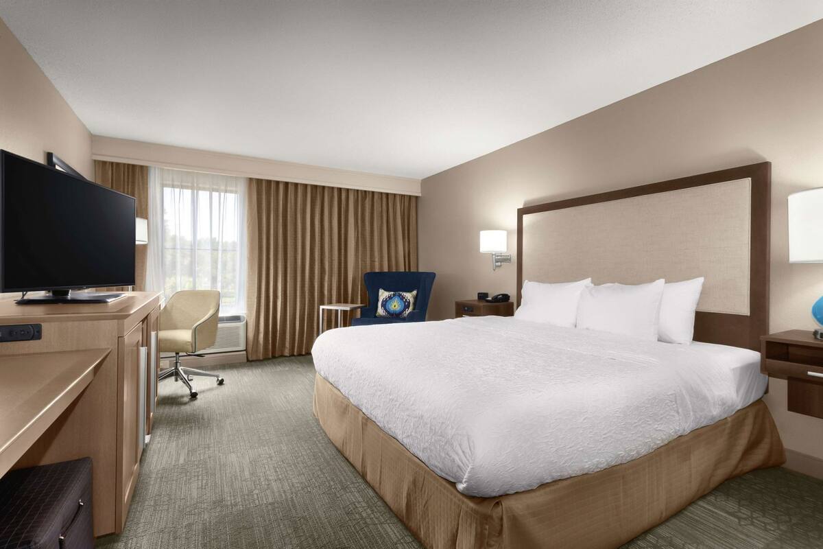 Una habitación de hotel con una cama grande, un escritorio con televisor, una silla, una mesita y una mesita de noche.