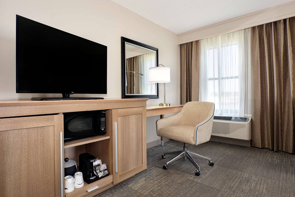 Une chambre d’hôtel équipée d’une télévision à écran plat, d’un bureau avec une lampe, d’une chaise, d’un four à micro-ondes, d’une machine à café et d’une fenêtre avec des rideaux.