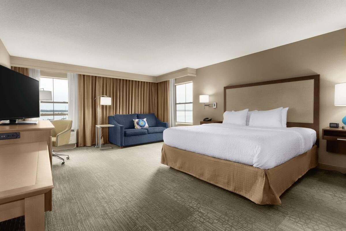 Habitación de hotel con una cama grande, un escritorio con televisor, un sofá y vistas al exterior.