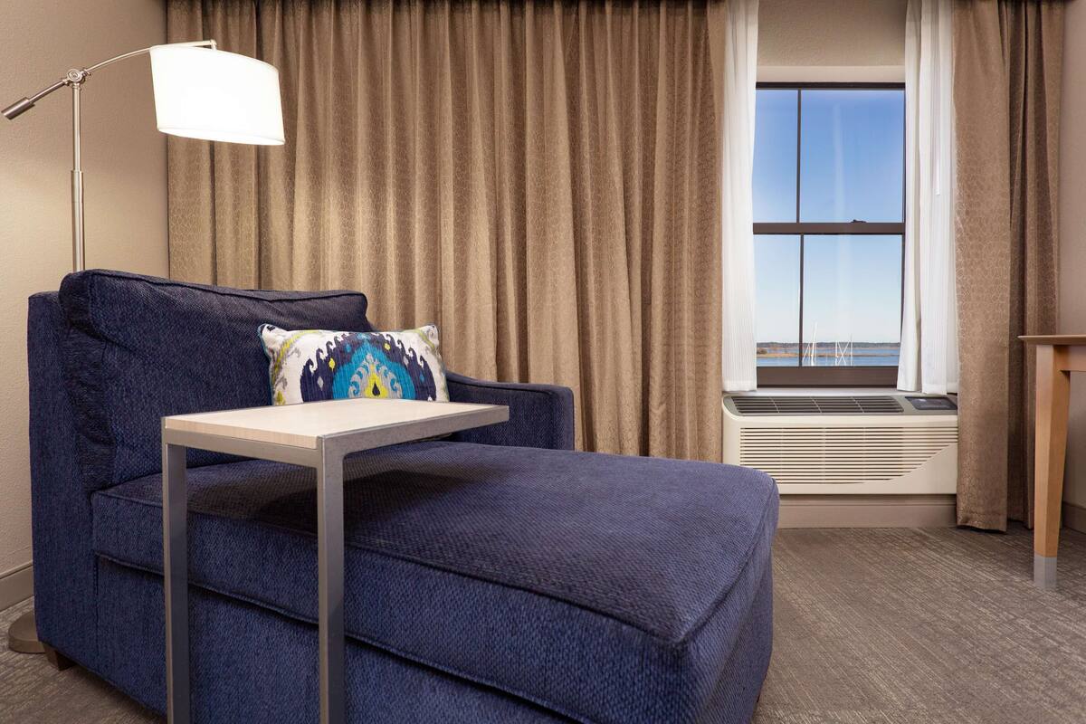 Une chambre d’hôtel avec un canapé bleu, une petite table, une fenêtre avec des rideaux et un radiateur.