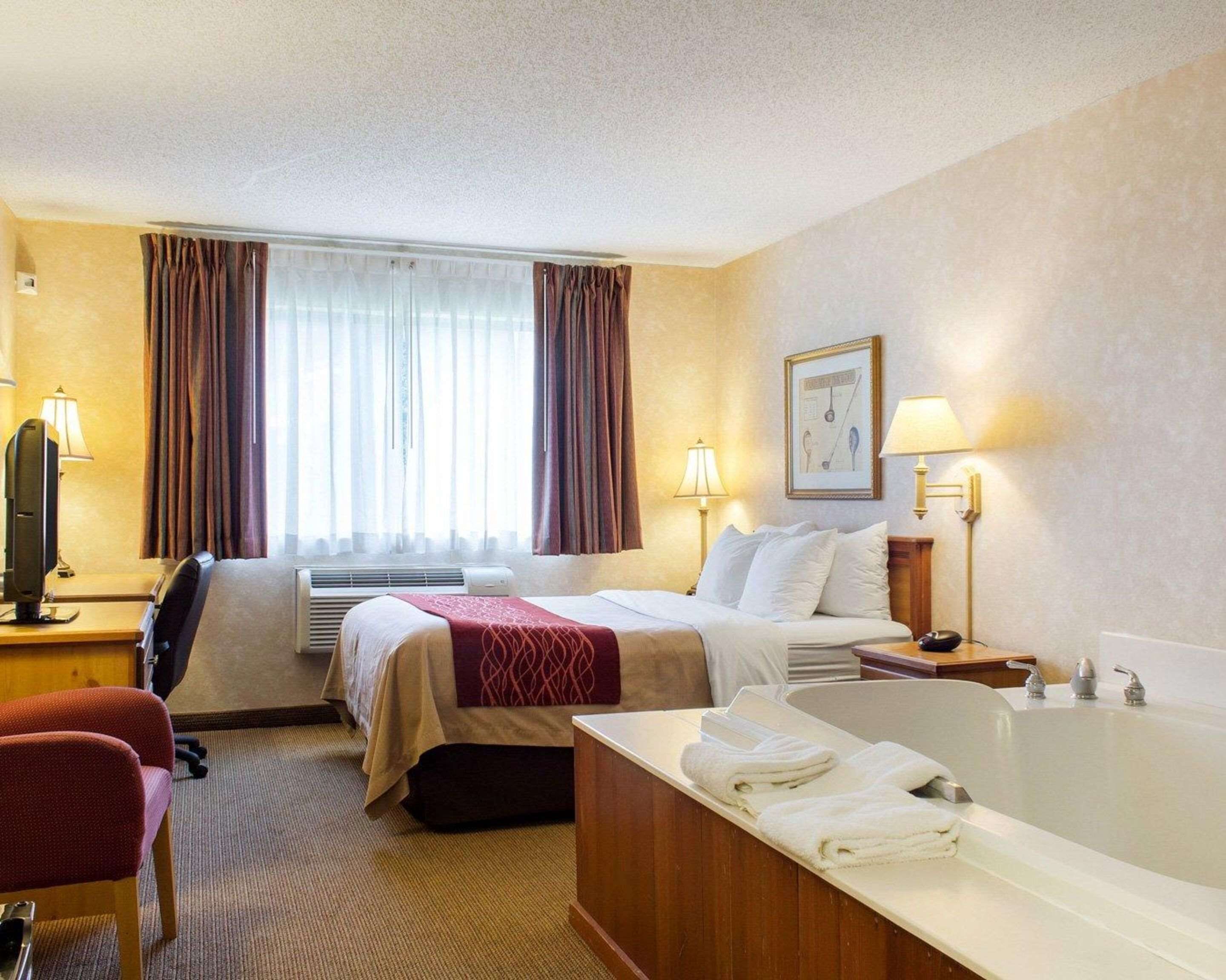 A hotel room with a bed, a desk with a computer, a chair, a desk lamp, a wall-mounted TV, a window with curtains, a wall-mounted picture, a bedside table with a lamp, and a bathtub with towels.
