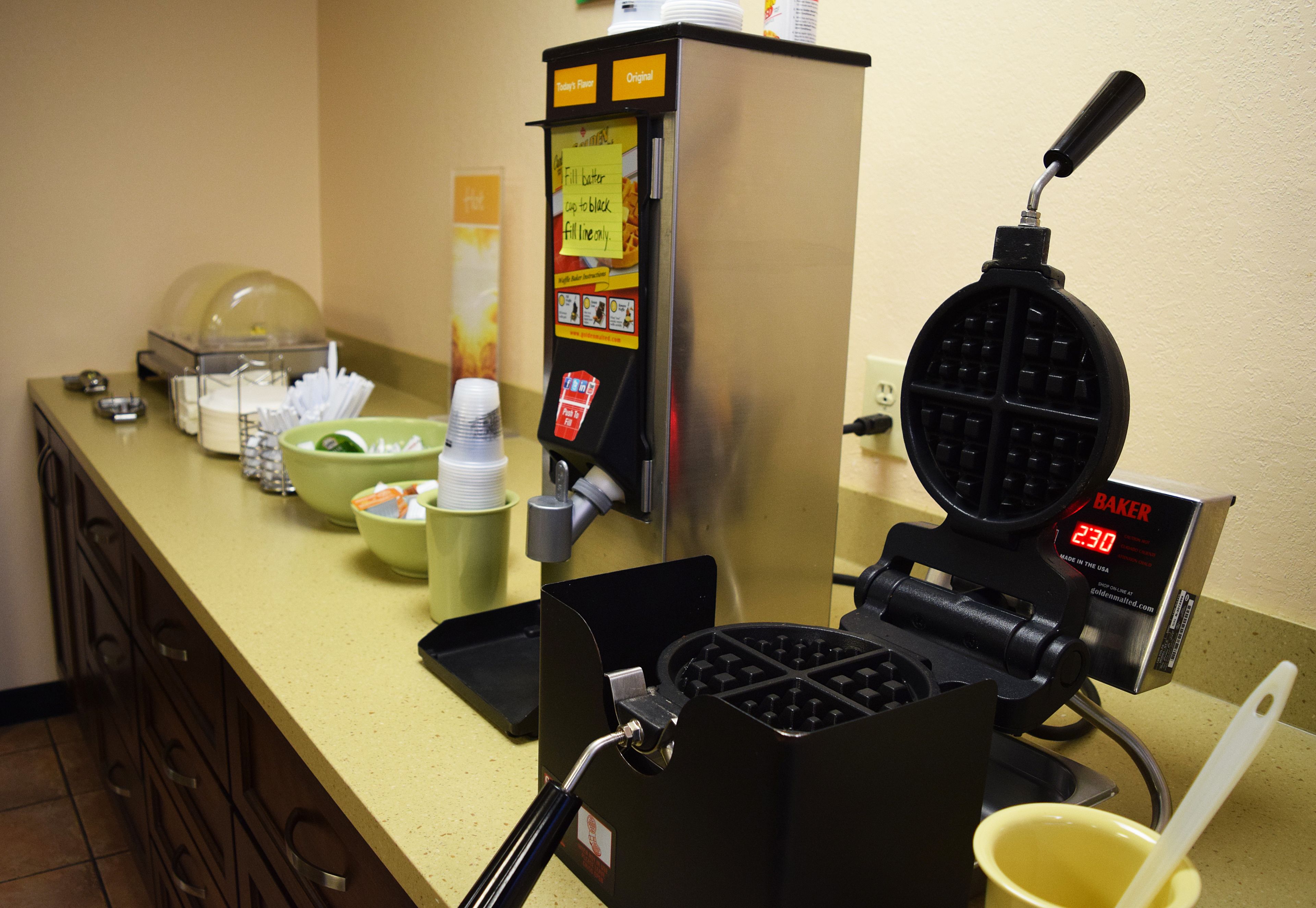 Una máquina para hacer gofres con pantalla digital, un bol de fruta y un dispensador de café sobre una encimera.