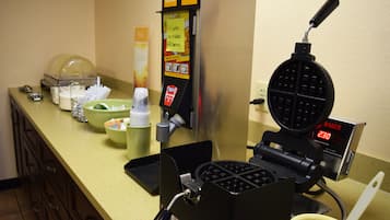 Una máquina para hacer gofres con pantalla digital, un bol de fruta y un dispensador de café sobre una encimera.
