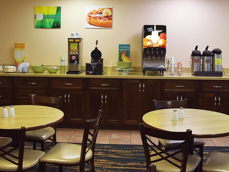 A hotel breakfast area with a buffet setup, including a coffee machine, fruit juice dispenser, and various food items. There are two round tables with chairs, and the room has wooden cabinets and a tiled floor.