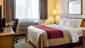 A hotel room with a bed, a desk, a chair, an air conditioner, a lamp, and a framed picture on the wall. - Quality Inn Brandon - Sioux Falls (Brandon)