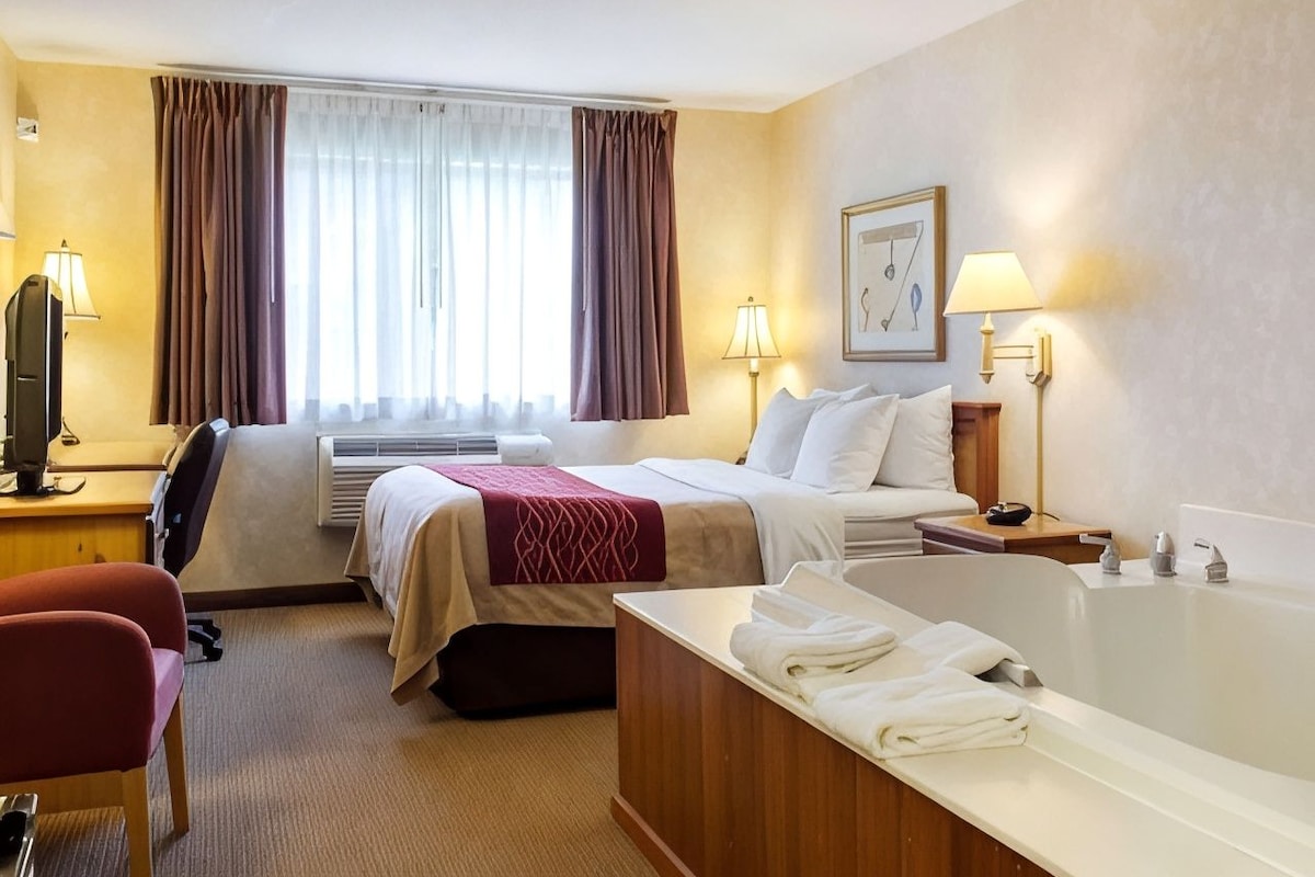 A hotel room with a bed, a desk with a computer, a chair, a bathtub, and a window with curtains.