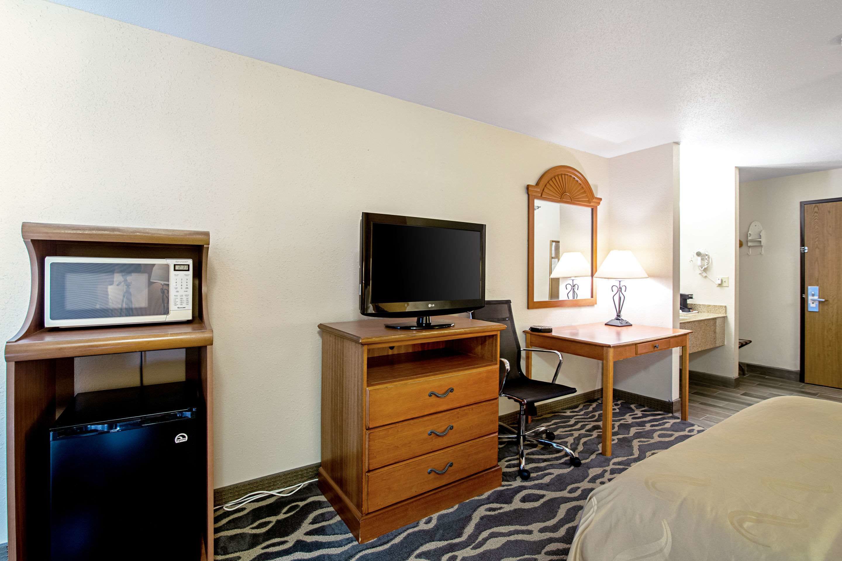 A hotel room with a bed, a desk with a television, a microwave, a chair, and a lamp.