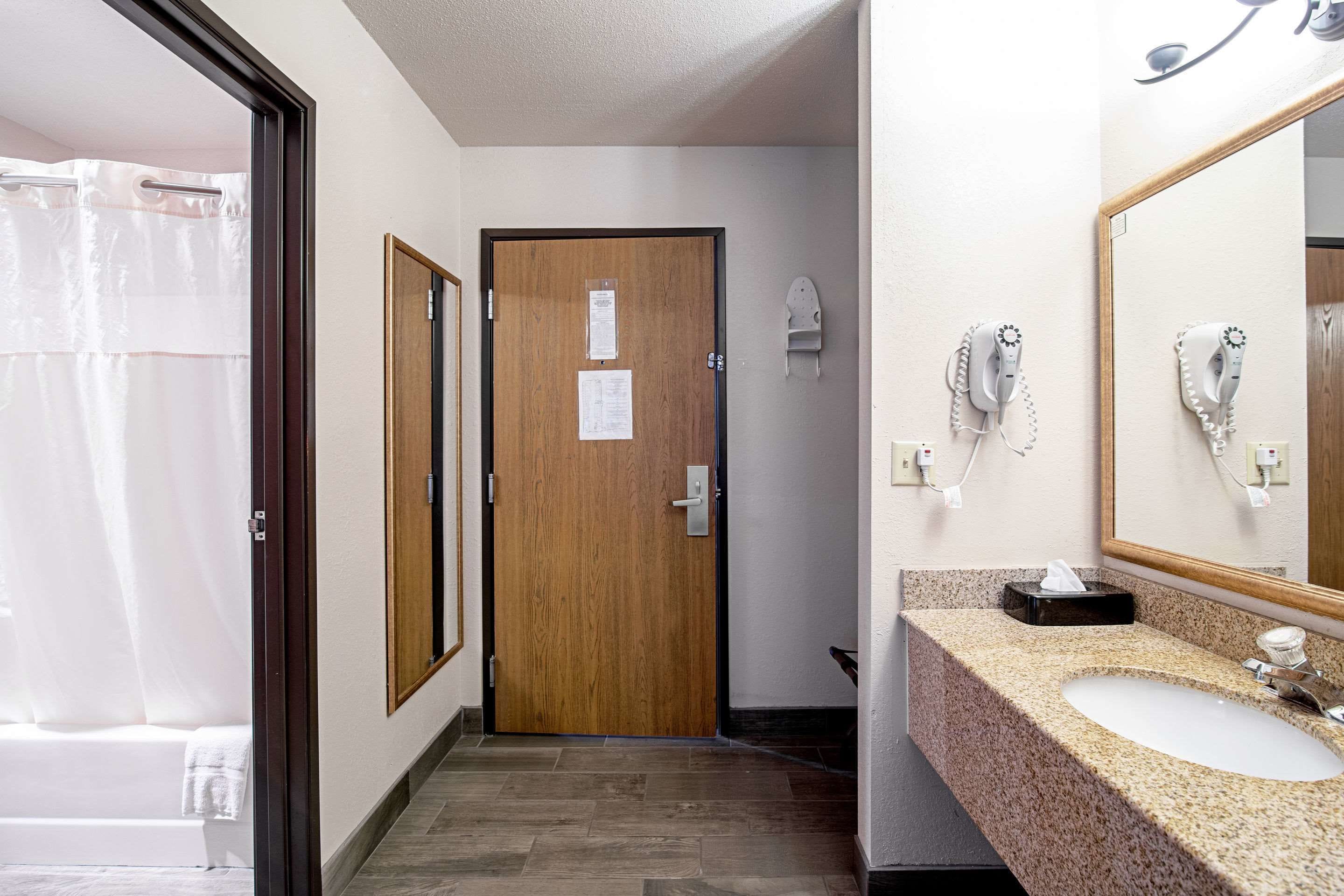 A hotel bathroom with a wooden door, a sink with a mirror, and a shower curtain.