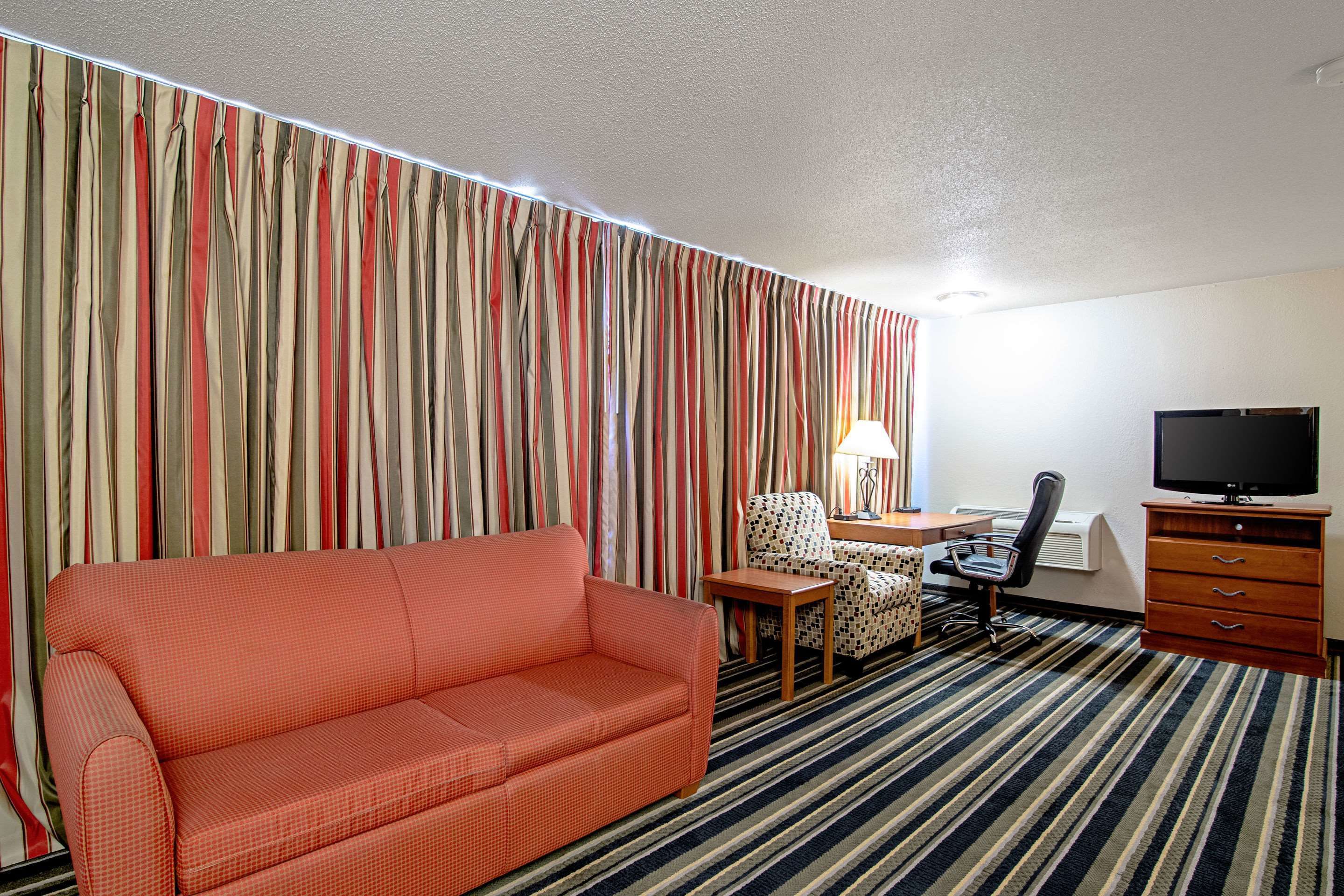 A hotel room with a striped curtain, a sofa, a chair, a desk with a lamp, a television, and a dresser.