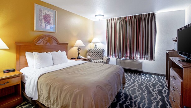A hotel room with a bed, two bedside tables with lamps, a patterned armchair, a television, and a window with curtains. - Quality Inn Huron (Huron)