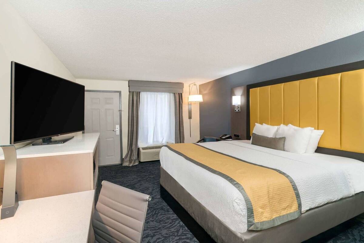 Habitación de hotel con cama, escritorio con televisor, silla, ventana con cortinas y lámpara de pared.