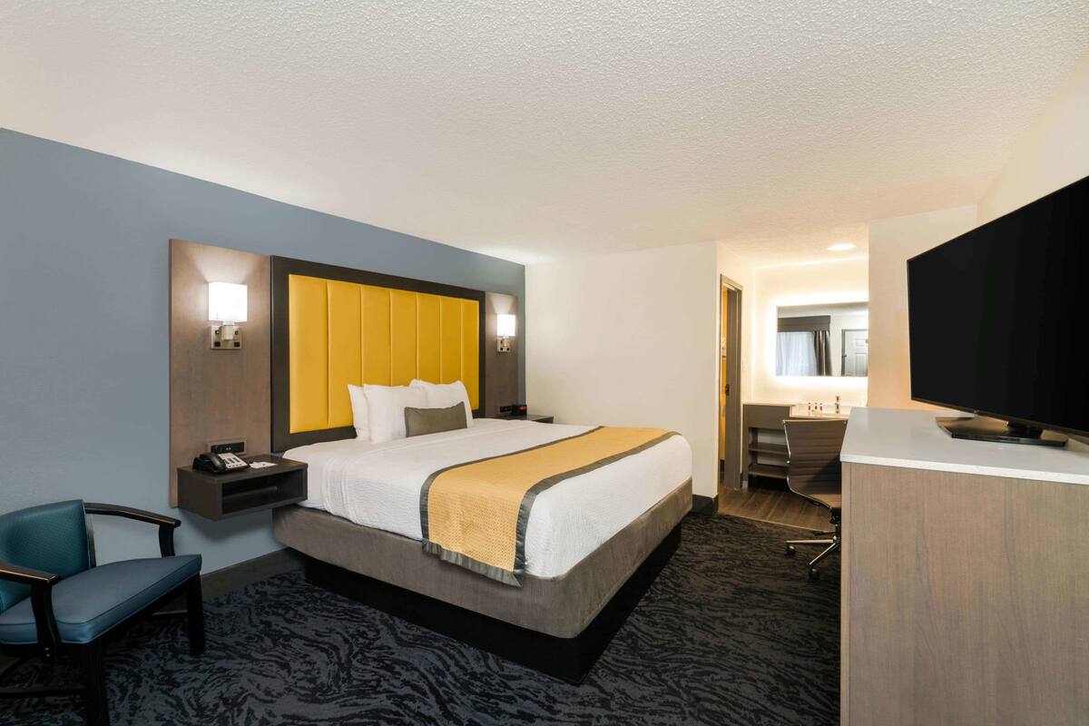 Habitación de hotel con cama, mesita de noche, silla, escritorio, televisor y baño visible a través de una puerta abierta.