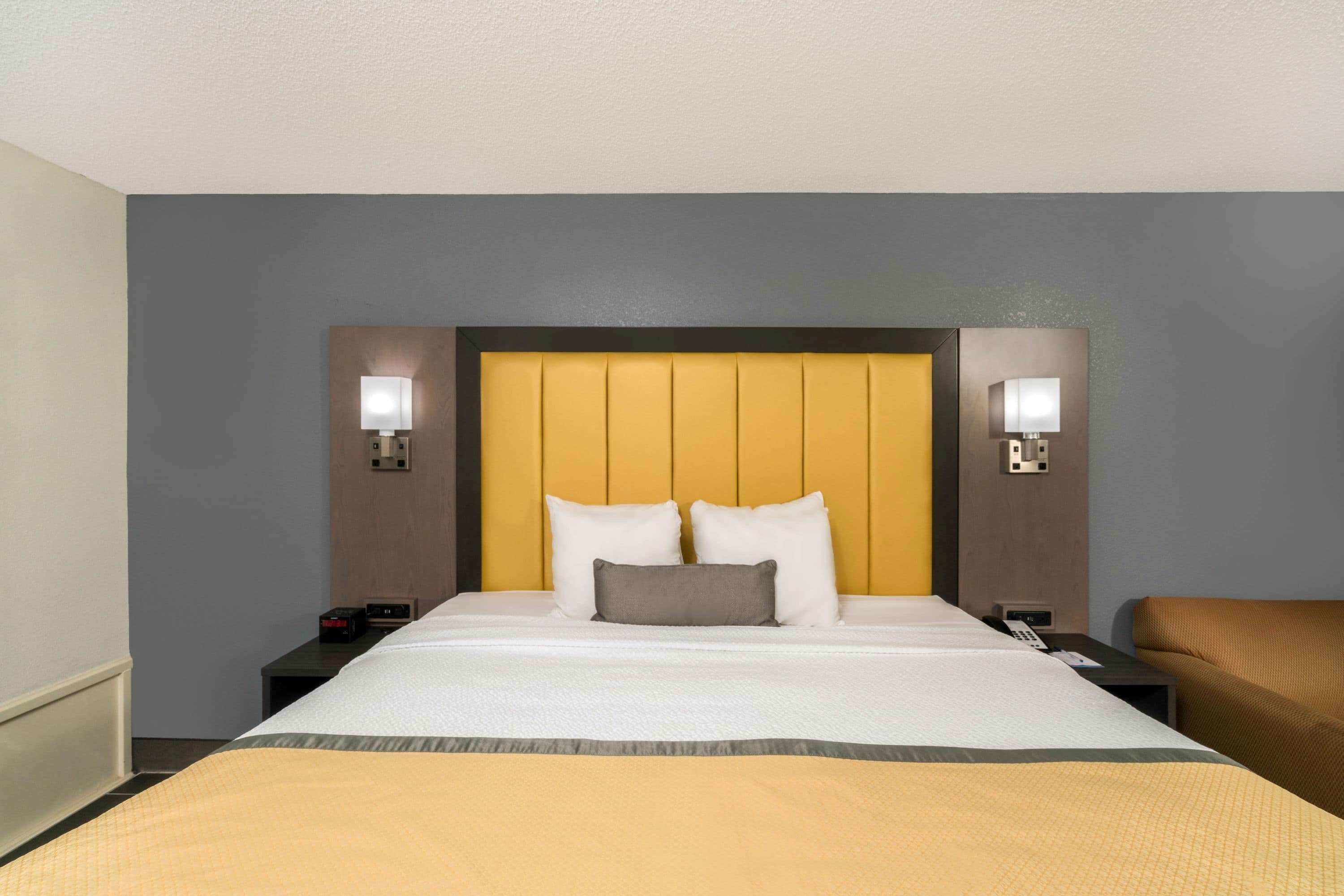 A hotel room with a large bed, two bedside tables, a nightstand, a chair, and a headboard with built-in lighting.