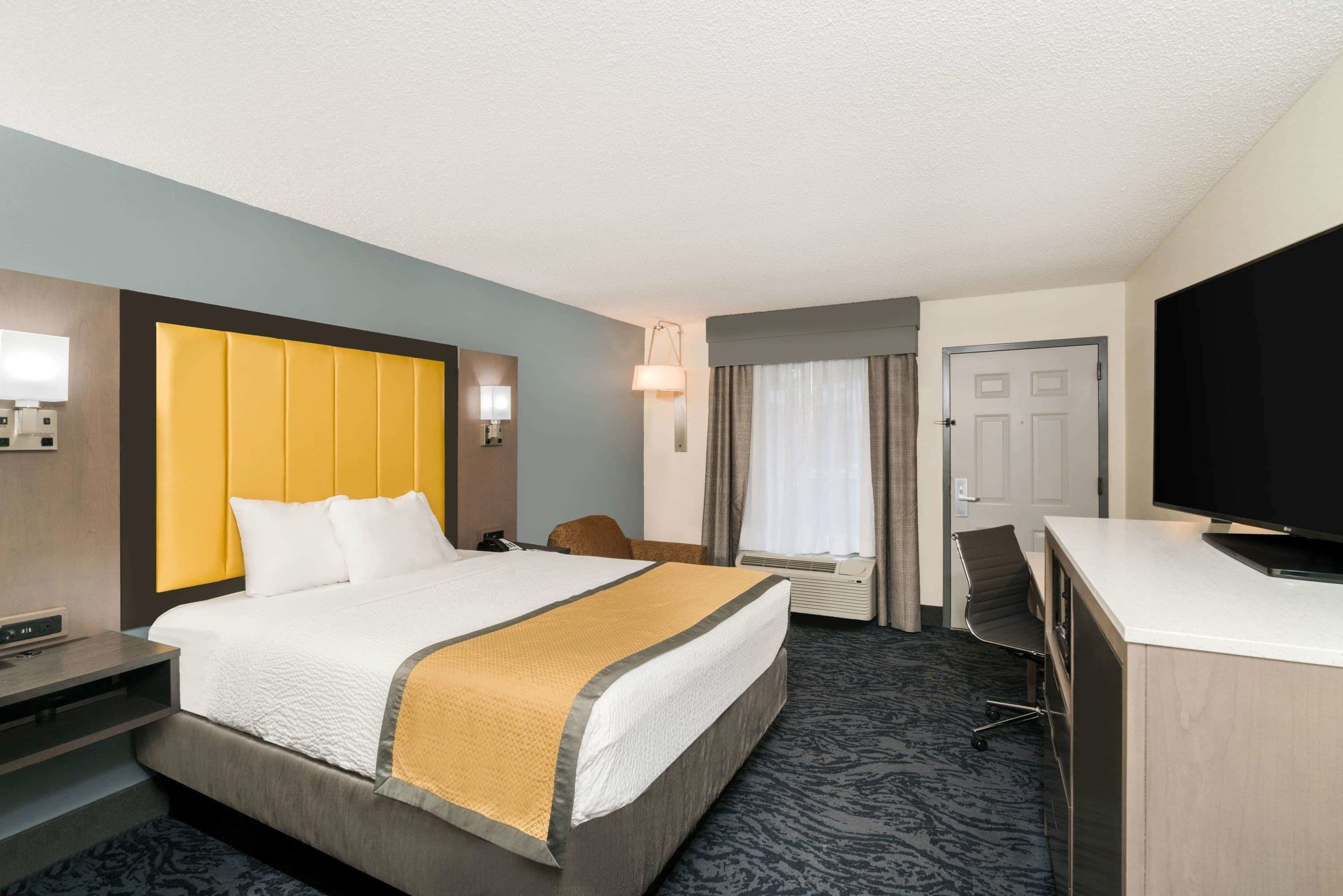 A hotel room with a large bed, a desk, a chair, a television, and a door.