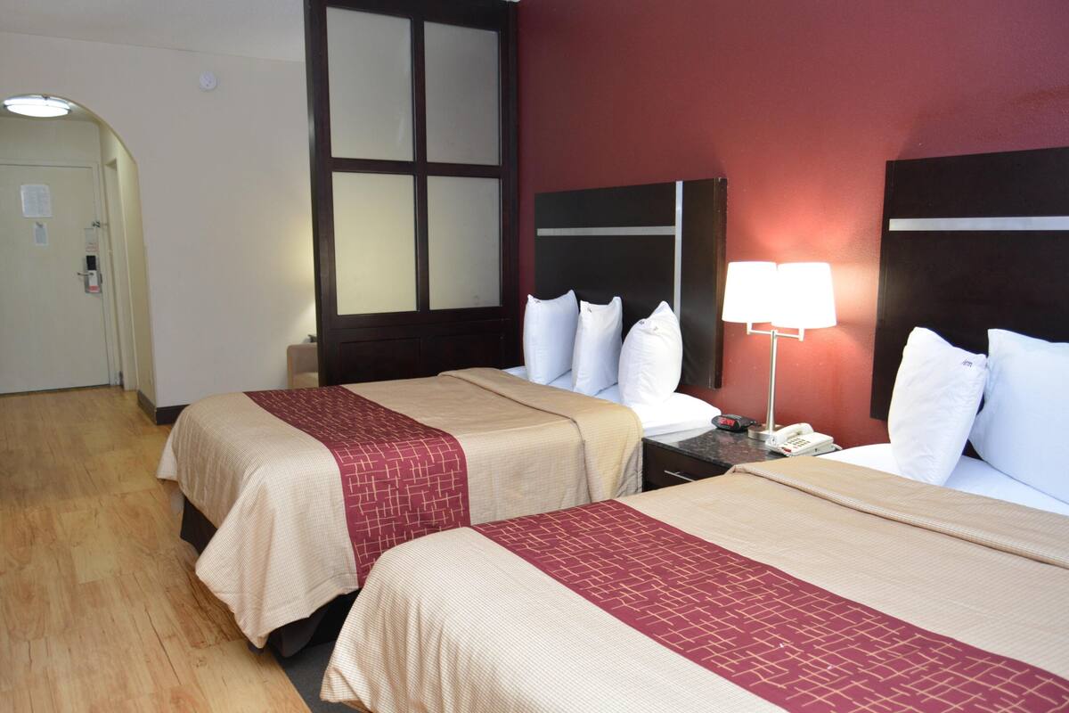 A hotel room with two beds, wooden flooring, a large window, and a nightstand with a lamp.