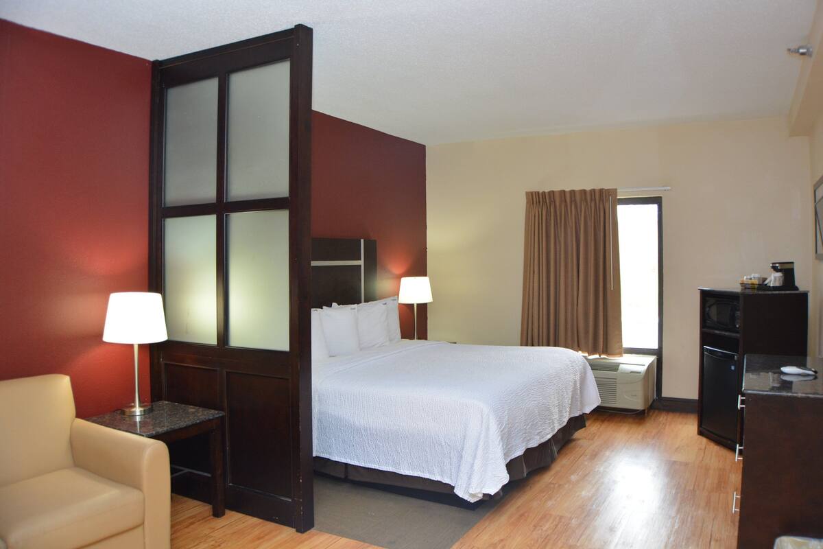 A hotel room with a bed, a desk, a chair, a window with curtains, and a wall with a frosted glass panel.