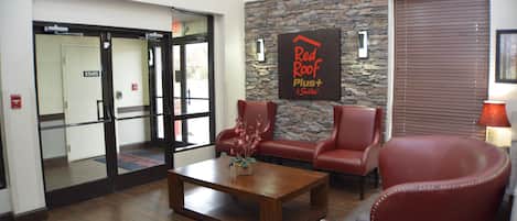 A lobby with a stone wall, a Red Roof Plus+ sign, leather chairs, a wooden coffee table, and an exit door.