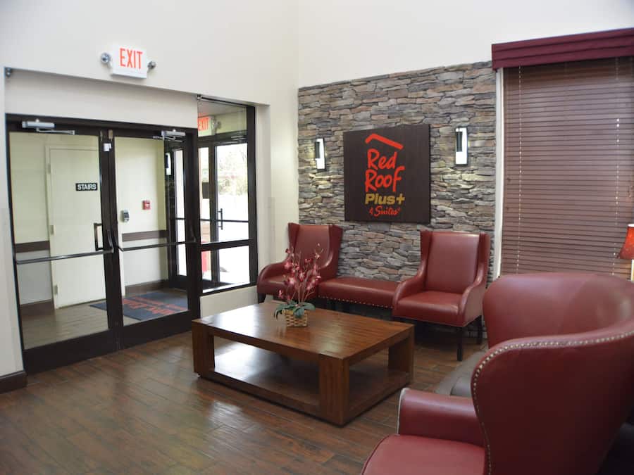 A lobby with a stone wall, a Red Roof Plus+ sign, leather chairs, a wooden coffee table, and an exit door.