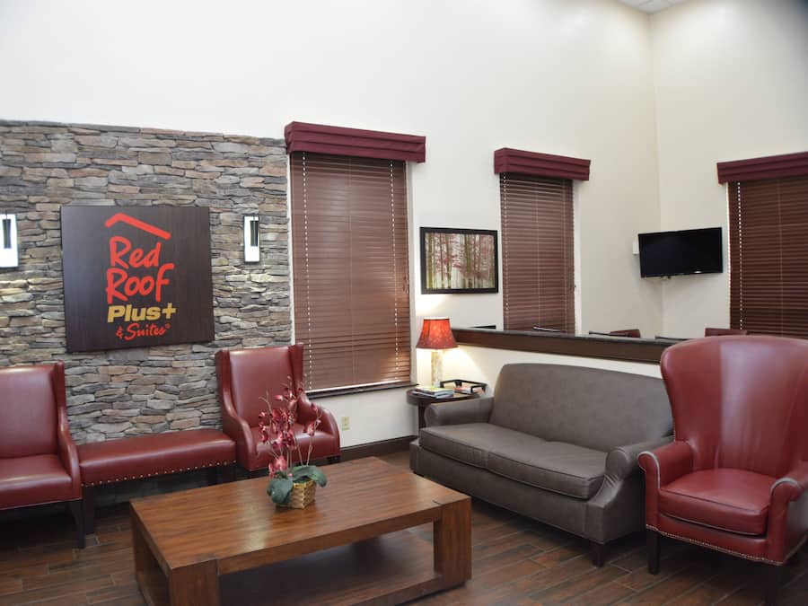 A furnished living area with a stone accent wall, a Red Roof Plus Suites sign, a flat-screen TV, and a coffee table with a floral arrangement.